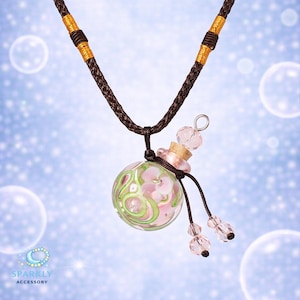 Könnte beinhalten: Halskette mit einem Glasanhänger in Form einer kleinen Flasche. Der Anhänger hat rosa Blumendesigns und grüne Akzente. Die Halskette hat eine braune Kordel mit goldenen Details und rosa Kristallperlen.