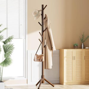 May include: A dark brown wooden coat rack with multiple branches for hanging items. A beige hat, coat, and purse are displayed. The rack has a tripod base and is positioned in a room with a window and cabinet.
