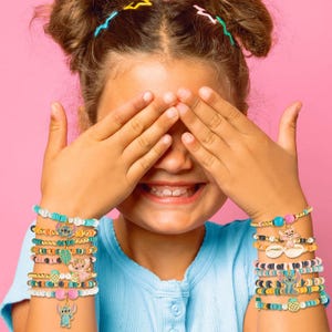 Puede incluir: Una persona joven con las manos cubriendo sus ojos, usando m&uacute;ltiples pulseras de cuentas. Las pulseras presentan varios colores, dijes y dise&ntilde;os, incluyendo personajes de dibujos animados y elementos tropicales. El fondo es rosa s&oacute;lido.