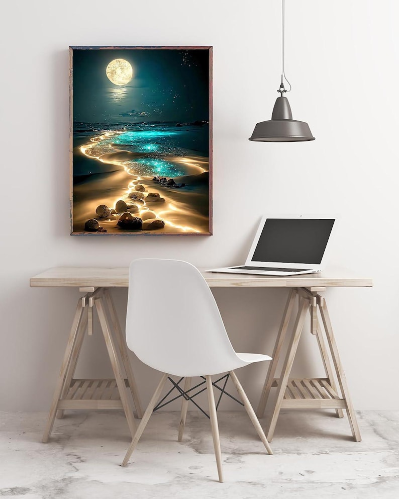 May include: A framed art print depicts a moonlit beach scene. The artwork features a bright full moon, a glowing path of light across the water, and a sandy shore with illuminated rocks. The print is displayed above a wooden desk with a laptop and a white chair.