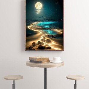 May include: A framed art print depicts a moonlit beach scene with a glowing path of light leading to the horizon. The artwork features a full moon, sparkling water, and a sandy shore. A small round table with books and a coffee cup is in front of the artwork.
