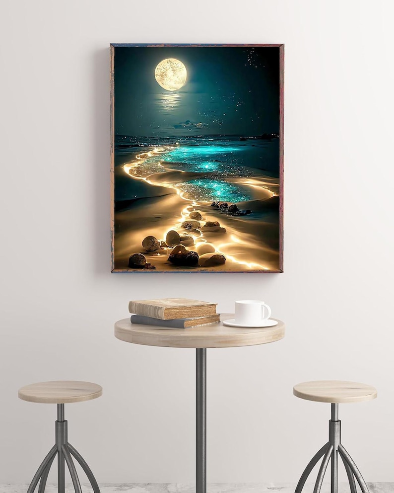 May include: A framed art print depicts a moonlit beach scene with a glowing path of light leading to the horizon. The artwork features a full moon, sparkling water, and a sandy shore. A small round table with books and a coffee cup is in front of the artwork.