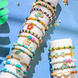 Puede incluir: Una colecci&oacute;n de pulseras de cuentas coloridas con varios dijes, incluyendo una flor roja, una mariposa, una hoja y un personaje de dibujos animados. Las pulseras se exhiben sobre una forma cil&iacute;ndrica blanca sobre un fondo azul.