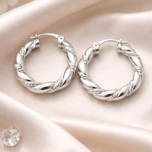 May include: Two silver hoop earrings with a twisted rope design. The earrings are presented on a cream satin fabric, with a small diamond-like gem and white flowers in the background. The earrings have a polished finish.