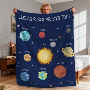 May include: A navy blue blanket featuring a solar system design with planets, stars, and the text "LUCAS'S SOLAR SYSTEM". Planets include Uranus, Mars, Mercury, Venus, Neptune, Earth, Jupiter, Saturn, and Pluto. The blanket is held up by a person.
