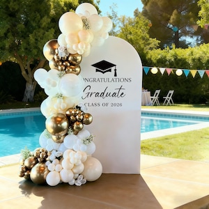 May include: A white graduation party backdrop with the text "Congratulations Graduate Class of 2026" and a mortarboard graphic. The backdrop is decorated with a balloon arch in white, gold, and cream colours. The scene is set outdoors near a swimming pool.