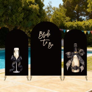 Puede incluir: Fondo negro arqueado con copa de champ&aacute;n, botella de champ&aacute;n y texto "Bride To Be". La copa y la botella tienen lazos negros. El tel&oacute;n de fondo est&aacute; montado cerca de una piscina.