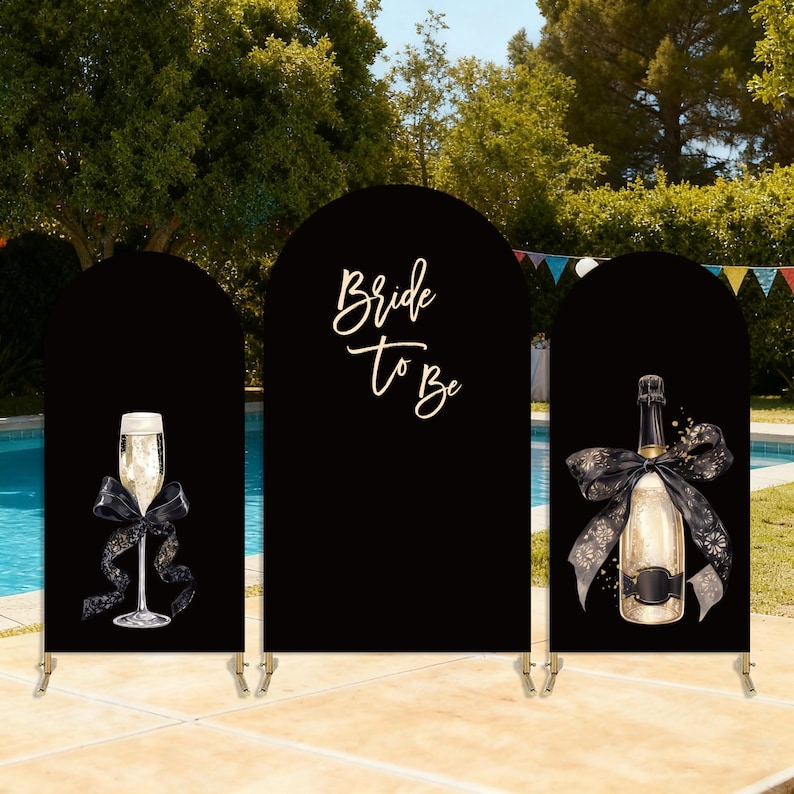Puede incluir: Fondo negro arqueado con copa de champ&aacute;n, botella de champ&aacute;n y texto "Bride To Be". La copa y la botella tienen lazos negros. El tel&oacute;n de fondo est&aacute; montado cerca de una piscina.