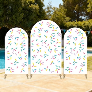 Puede incluir: Tres paneles de fondo blancos arqueados con diseños de chispas coloridas. Los paneles están decorados con chispas azules, amarillas, rosas y verdes. Los paneles están instalados al aire libre en un patio cerca de una piscina.