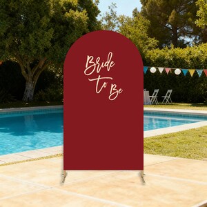 Puede incluir: Un letrero en forma de arco de color burdeos con las palabras "Bride To Be" en una elegante escritura. El letrero est&aacute; sostenido por un soporte de metal dorado y est&aacute; instalado al aire libre cerca de una piscina, con una pancarta festiva en el fondo.