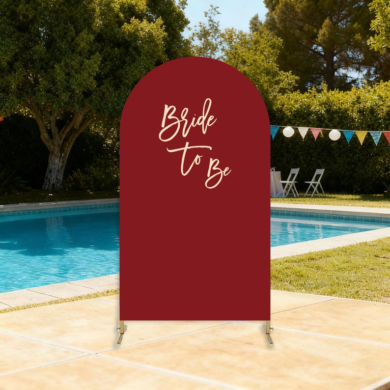 Puede incluir: Un letrero en forma de arco de color burdeos con las palabras "Bride To Be" en una elegante escritura. El letrero est&aacute; sostenido por un soporte de metal dorado y est&aacute; instalado al aire libre cerca de una piscina, con una pancarta festiva en el fondo.