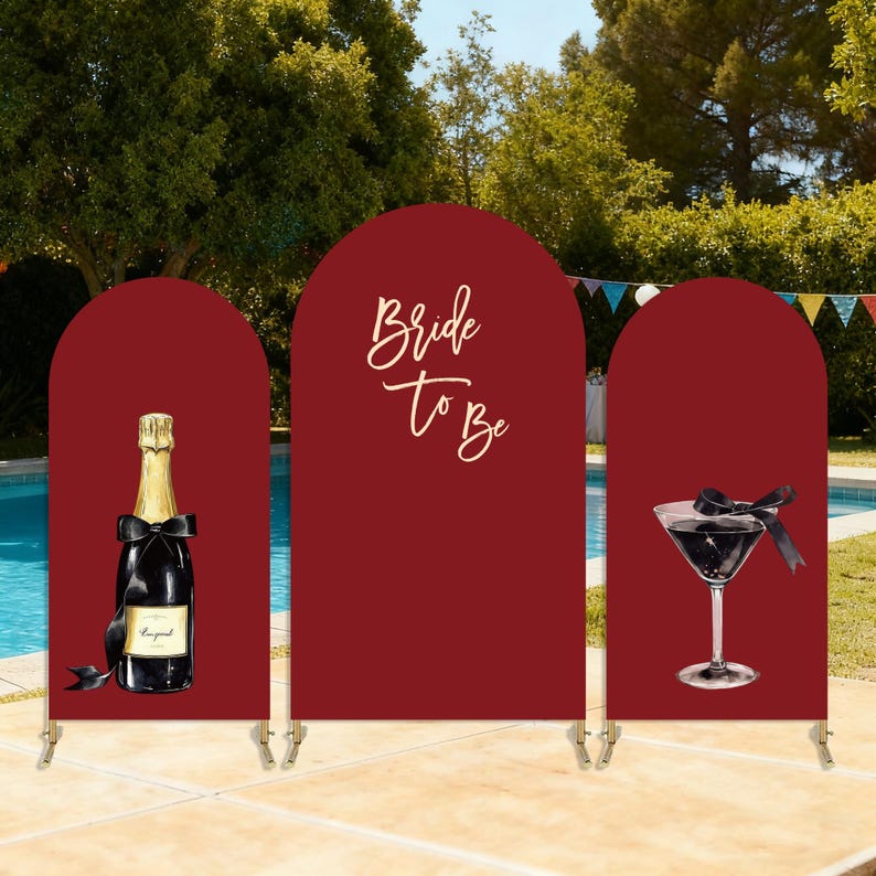 Puede incluir: Un tel&oacute;n de fondo burdeos con tres paneles arqueados. Un panel presenta una botella de champ&aacute;n con un lazo negro, otro el texto "Bride To Be", y el &uacute;ltimo muestra una copa de martini con un lazo negro. El tel&oacute;n de fondo est&aacute; instalado al aire libre.