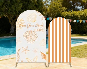 May include: Two arched backdrop panels. One features a cream background with gold seashell illustrations and the text "Sea You Soon BABY." The other panel has vertical orange and white stripes. Both are supported by metal stands.