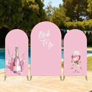 Può includere: Uno sfondo rosa a tre pannelli con sommità ad arco. Il pannello di sinistra presenta una bottiglia di champagne con effetto splash. Il pannello centrale ha il testo "Bride To Be". Il pannello destro mostra un calice di vino con un nastro dorato.
