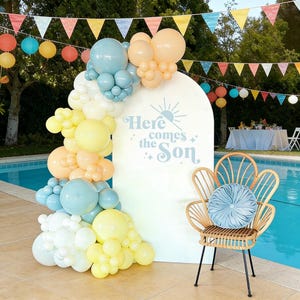 Puede incluir: Una escena de fiesta con un arco blanco decorado con globos pastel en amarillo, azul y melocotón. El arco tiene el texto "Here comes the Son". Una silla de mimbre con un cojín azul está cerca. Guirnaldas y faroles de papel de colores decoran el fondo.