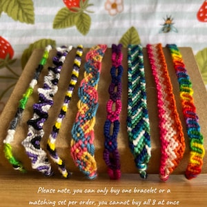 May include: Eight handmade friendship bracelets in various colours, including green, purple, yellow, pink, blue, orange, and rainbow. Each bracelet has a distinct woven or braided pattern. The bracelets are presented on a brown surface.