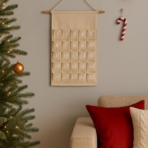 Peut inclure: Calendrier de l'Avent en tissu beige avec 25 poches numérotées, suspendu à une tringle en bois. Un sapin de Noël, des ornements et un sucre d'orge sont également visibles.