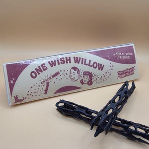May include: A rectangular box labeled ONE WISH WILLOW with illustrations of faces, stars, and the text AMAZE YOUR FRIENDS and You only get ONE WISH. Two black, cylindrical, porous, and textured objects are positioned in front of the box.