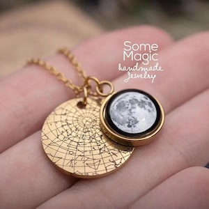 May include: A gold-colored necklace with two pendants. One pendant is a circular disc with a detailed star map design. The other pendant is a round, black-rimmed charm with a detailed image of the moon. The text "Some Magic handmade jewelry" is in the background.