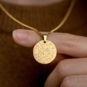 May include: A gold-colored necklace with a round pendant featuring a constellation design. The pendant hangs from a delicate chain. The necklace is held by a person.