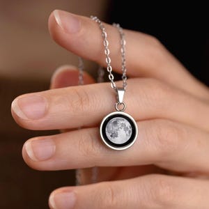 May include: Silver-toned necklace featuring a circular pendant with a detailed image of the moon. The pendant is framed in black and hangs from a delicate chain. A celestial-themed jewelry item.