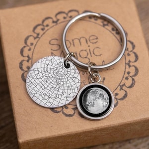 May include: A silver keychain with two charms. One charm is a round, detailed depiction of the moon. The other is a circular charm with a star map design. The keychain is resting on a brown cardboard box with the words "Some Magic" printed on it.