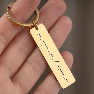 May include: A gold-colored rectangular keychain with a Morse code pattern engraved on it. The keychain has a circular ring attached to the top. The Morse code is in black and is engraved on the gold-colored metal.