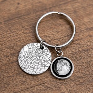 May include: Silver keychain with two charms. One charm is a round disc with a star map design. The other charm is a circular pendant with a detailed image of the moon. The keychain is attached to a silver key ring.
