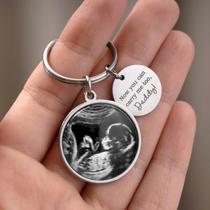 May include: A silver keychain with two charms. One charm is a circular pendant with a black and white sonogram image. The second charm is a small, round tag with the text "Now you can carry me too, Daddy!"