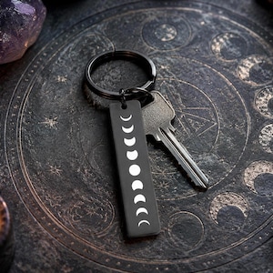 May include: Black rectangular keychain featuring a white moon phase design. The keychain is attached to a black ring with a silver key. The background is a dark, textured surface with celestial patterns and a lit candle.