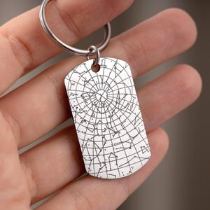 May include: A silver-colored dog tag-shaped keychain pendant. The pendant features a detailed etched design of a star map, with lines and dots representing constellations. The keychain ring is silver.