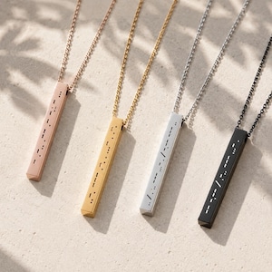 May include: Four pendant necklaces in rose gold, gold, silver, and black. Each necklace features a rectangular pendant with a Morse code design. The pendants hang from delicate chain necklaces, displayed on a light beige surface.