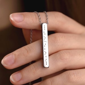 May include: A silver-colored necklace with a rectangular pendant. The pendant has a white surface with black dots and dashes, possibly Morse code. The necklace is held by a person.