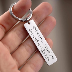 May include: A rectangular silver keychain with a ring, featuring the engraved message "Drive safe... I need you here with me Kim" and two heart symbols. The keychain is held in a hand.