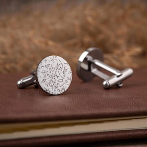 May include: A pair of silver cufflinks with a white face featuring a black abstract design. The cufflinks are resting on a brown leather-bound book, with a blurred background of dried plants.