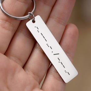 May include: A rectangular silver keychain tag with Morse code etched on it. The tag is attached to a silver keyring. The Morse code consists of dots and dashes. The background is blurred.
