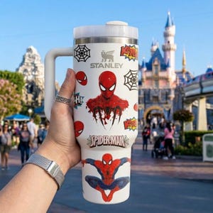 May include: White Stanley travel mug featuring Spider-Man graphics. The mug has a white handle and lid, with various Spider-Man images and the text "Spider-Man". The background shows a theme park with a castle.