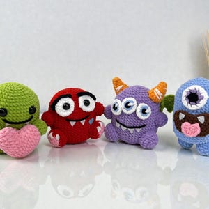 May include: A row of four handmade crochet monster toys. The monsters are in green, red, purple, and blue. The green monster holds a pink heart. The red monster has large eyes and a toothy grin. The purple monster has three eyes and horns. The blue monster has one eye and a wide mouth.