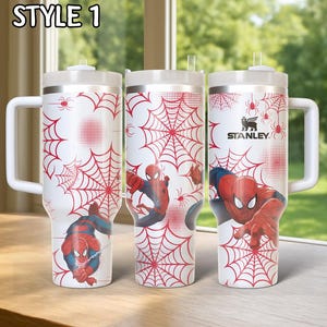 May include: Three white insulated tumblers with handles, featuring a red spiderweb design and illustrations of Spider-Man. The tumblers have a silver rim and lid, with the text "STYLE 1" at the top. The Stanley logo is visible on one tumbler.