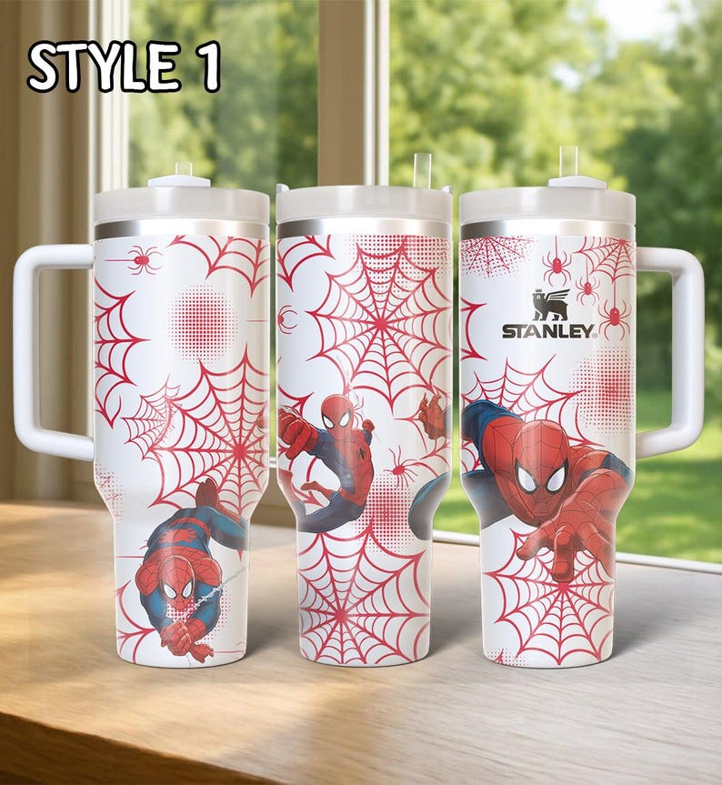 May include: Three white insulated tumblers with handles, featuring a red spiderweb design and illustrations of Spider-Man. The tumblers have a silver rim and lid, with the text "STYLE 1" at the top. The Stanley logo is visible on one tumbler.