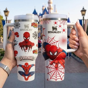 May include: Two white Stanley travel mugs featuring Spider-Man graphics. One mug has a red Spider-Man design with a web, the other has multiple Spider-Man images and the words "Spider-Man". Both mugs have white handles and lids.