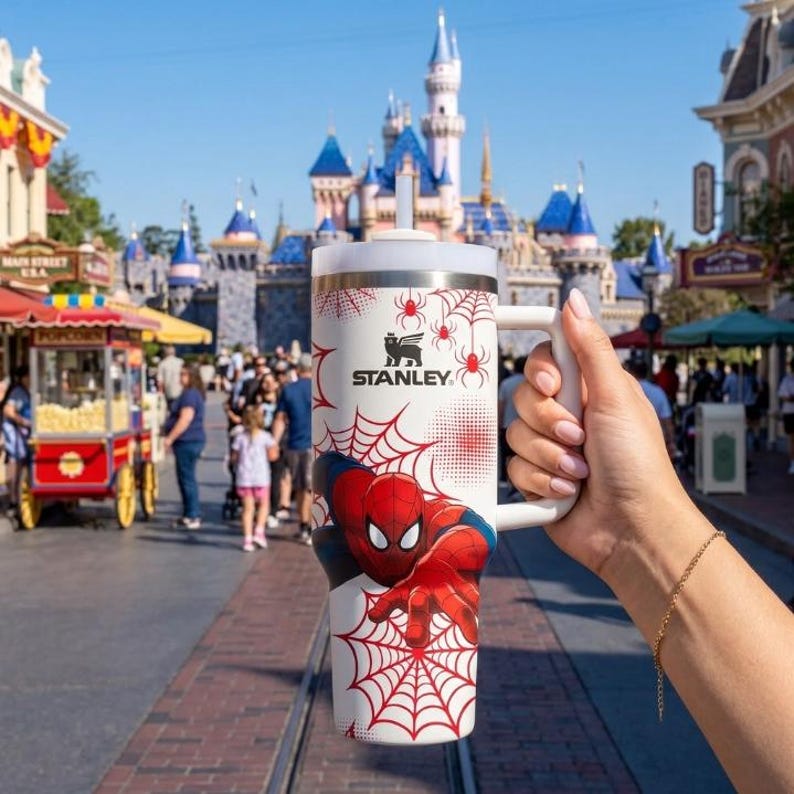 May include: A white Stanley travel mug featuring Spider-Man graphics and a red spiderweb design. The mug has a white handle and a silver rim. The Stanley logo is visible. The background shows a theme park with a castle.