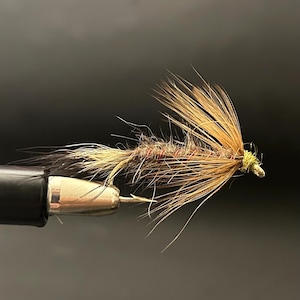 May include: Close-up of a handmade fly fishing lure. The lure features a hook with a brown body and tan feather accents. The fly is tied with various materials, including brown and tan feathers, and a touch of red thread.