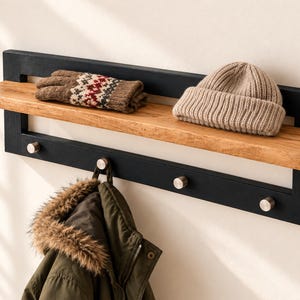 Puede incluir: Un perchero de pared con marco rectangular negro, estante de madera natural y cuatro ganchos de metal plateado. Sobre el estante hay guantes estampados y un gorro de punto beige, mientras que una chaqueta verde oliva cuelga debajo.
