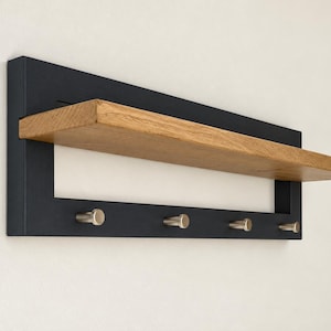 Puede incluir: Un estante de pared con un marco rectangular negro, una superficie de madera natural en tono marrón claro y cuatro pequeños ganchos cilíndricos de color plateado fijados en la barra horizontal inferior.