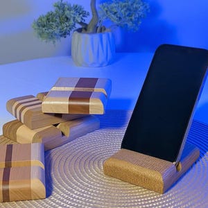 Peut inclure: Plusieurs supports de téléphone en bois. L'un d'eux maintient un téléphone à écran noir. D'autres supports sont empilés, présentant différents motifs de bois, notamment des rayures et des carreaux. Les supports sont faits de tons de bois clairs et foncés.