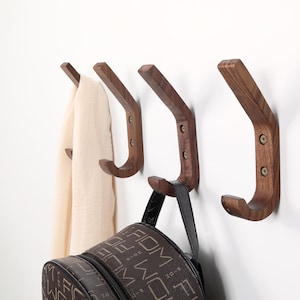 Puede incluir: Cuatro percheros de madera marrón oscuro fijados a una pared blanca. Los percheros tienen un diseño curvo, con una bufanda beige y una mochila marrón colgadas. Los percheros están asegurados con tornillos.