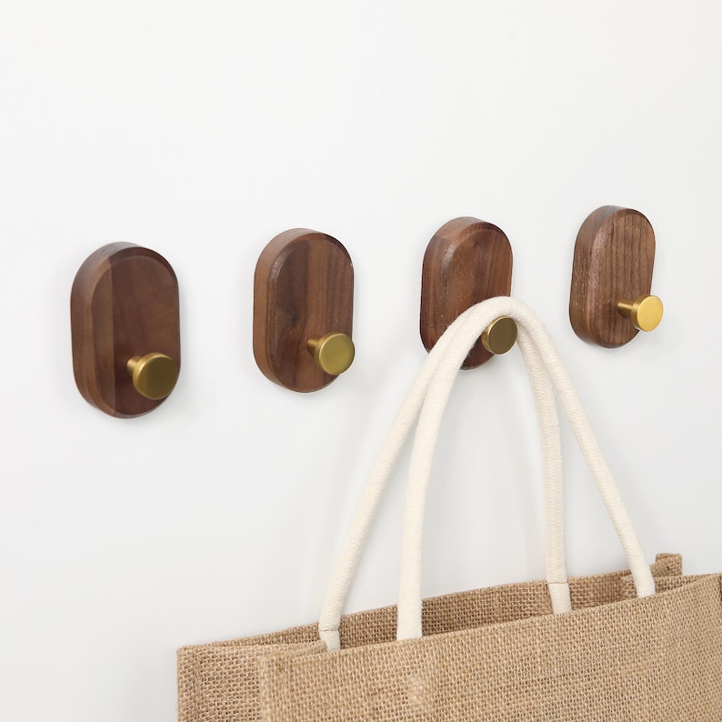 May include: Four wooden wall hooks with brass knobs are mounted on a white wall. The hooks are oval-shaped and made of dark wood. A beige tote bag with white rope handles hangs from one of the hooks.