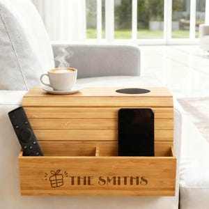 May include: A bamboo sofa arm tray with a cup of coffee, a remote control, and a smartphone. The tray has a built-in cup holder and a compartment with the engraved text "THE SMITHS" and a gift icon.