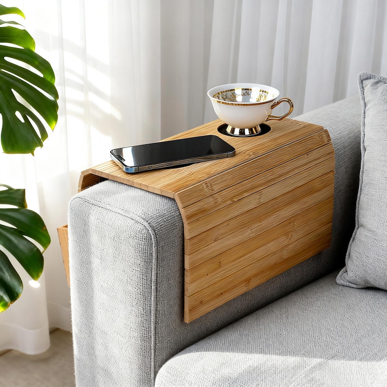 May include: A bamboo sofa arm tray with a built-in cup holder and a slot for a smartphone. A white teacup with gold trim sits in the holder. A black smartphone rests on the tray. The tray is placed on a gray sofa arm.